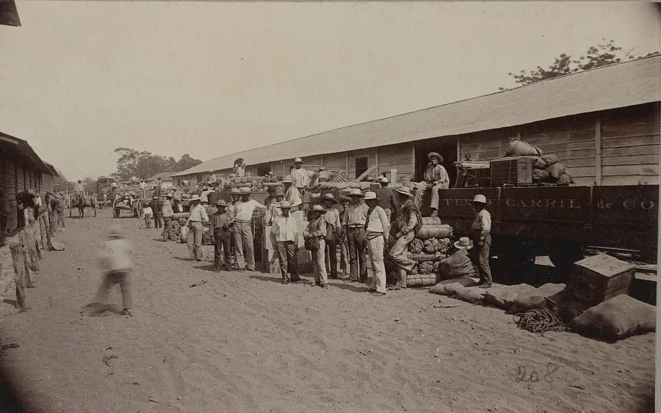 Estación del ferrocarril en Puntarenas hacia 1880, con hombres y carga amontonada junto a un vagón que lleva pintado en español el nombre del ferrocarril. Por este puerto del Pacífico desembarcaron en 1873 las 653 personas llegadas de Macao, contratadas para la misma obra que trajo a los jamaiquinos por el lado del Caribe: el ferrocarril al Atlántico. Fotografía de la Biblioteca del Congreso de Estados Unidos, dominio público.