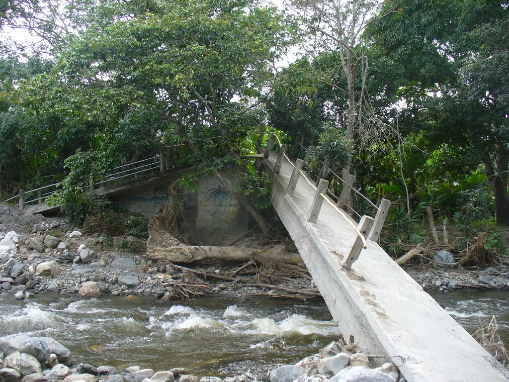 Un puente peatonal vencido sobre el río, con los troncos y la palizada que bajaron con la creciente amontonados contra la base. En el ejemplo de la lección, un puente cerrado es la causa de que el bus llegue tarde y la ruta vieja es apenas el paso intermedio. Por eso, cuando el enunciado pide el motivo, hay que seguir la cadena hasta donde el texto la arma. Imagen de Flickr, betoscopio, licencia CC BY-SA.