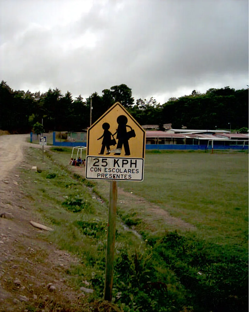 Una señal preventiva de zona escolar en una carretera de Costa Rica, con el límite de 25 km/h. Las señales preventivas advierten un peligro adelante; no obligan como las reglamentarias ni orientan como las de información. Fotografía de Flickr, Daquella manera, licencia CC0.