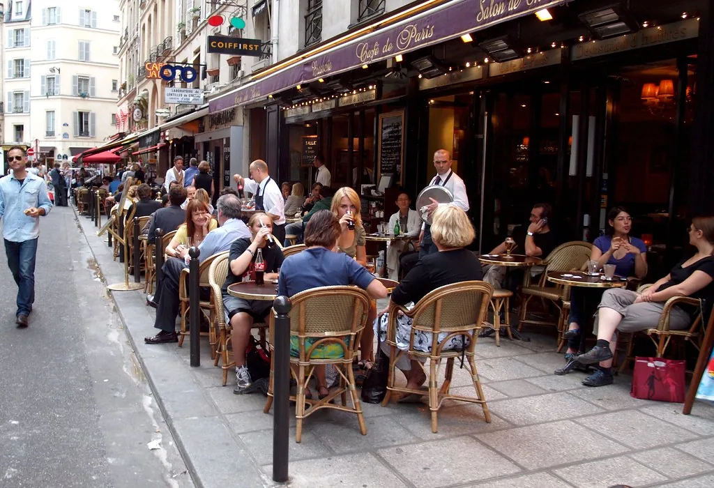 La terraza de un café en una calle de Francia, con la gente conversando en las mesas. Sentarse ahí a conversar es justo un hábito colectivo de los que nombra la lección, el tipo de costumbre que en francés se cuenta con on y no con un sujeto en particular. Imagen de flickr, zoetnet, licencia BY.