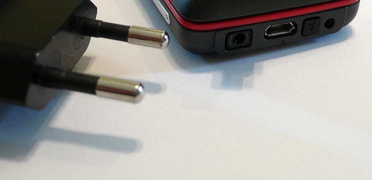 Un cargador junto a un celular. Plug in the charger and unplug it when the battery is full. Foto: Reinraum, Wikimedia Commons, dominio público (recortada).