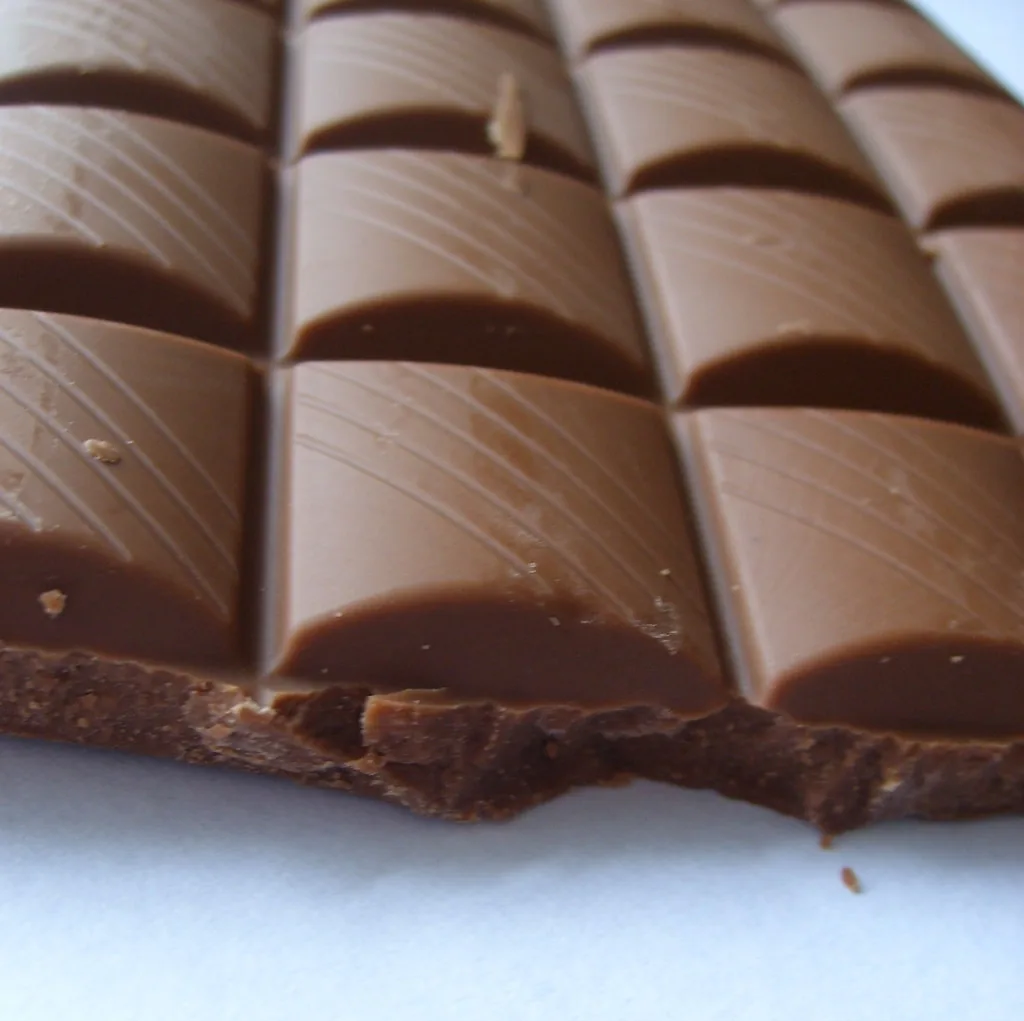 Una barra de chocolate marcada en cuadros. Las rayas a lo largo y las rayas a lo ancho parten la barra dos veces, y por eso los cuadritos que quedan son tantos como el producto de las dos particiones. Al multiplicar fracciones pasa lo mismo: se parten las partes, y por eso los denominadores se multiplican. Imagen de flickr — Siona Karen, licencia BY.