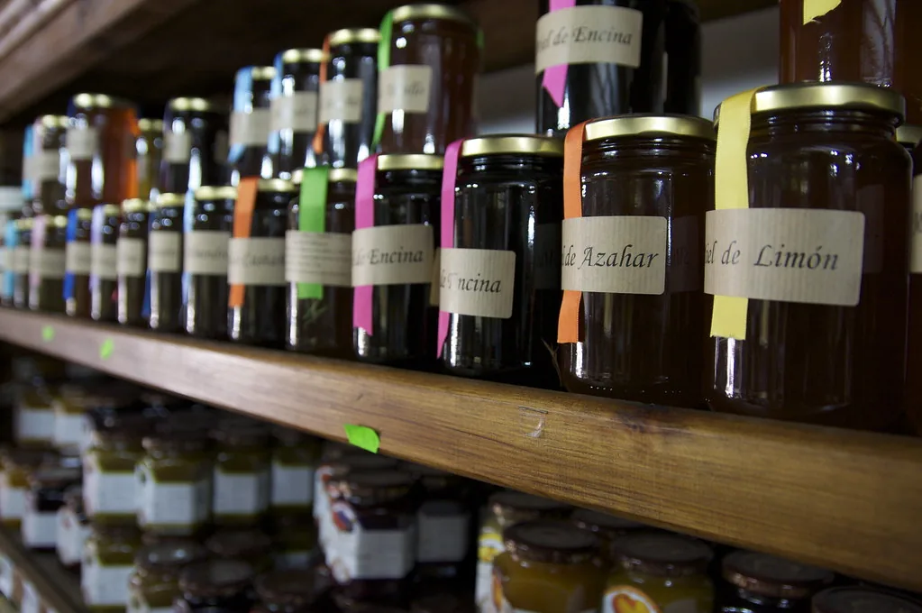 Frascos de miel en el estante de una venta, cada uno con su etiqueta. Si la miel se envasa en frascos de 0,8 litros, con 40 litros se llenan 50 frascos: se dividió y el resultado salió mayor que el número de partida. Eso ocurre siempre que se divide entre un número menor que uno, porque lo pequeño cabe muchas veces. Imagen de flickr — psmithy, licencia BY.